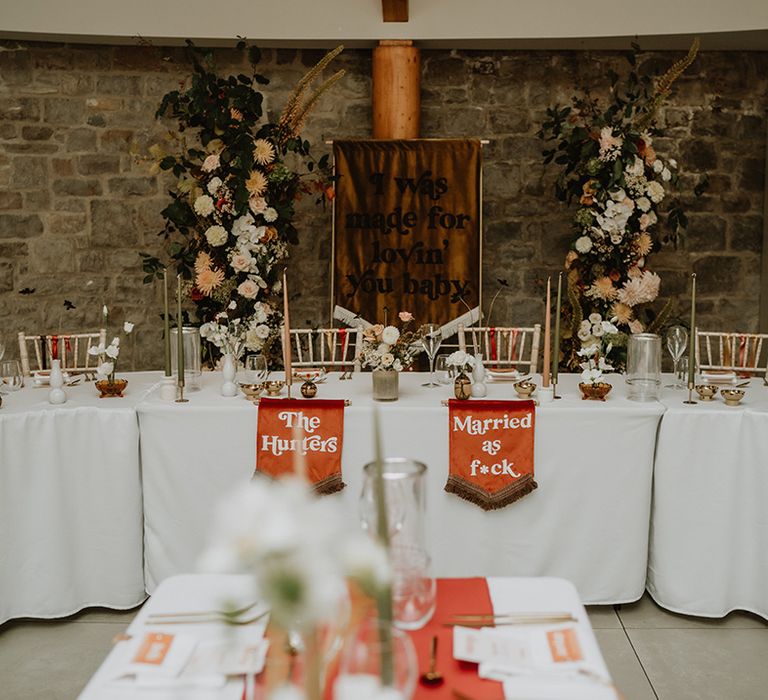 70's inspired wedding tablescape with burnt orange backdrop banner, two orange fabric wedding signs and large floral decoration pillars