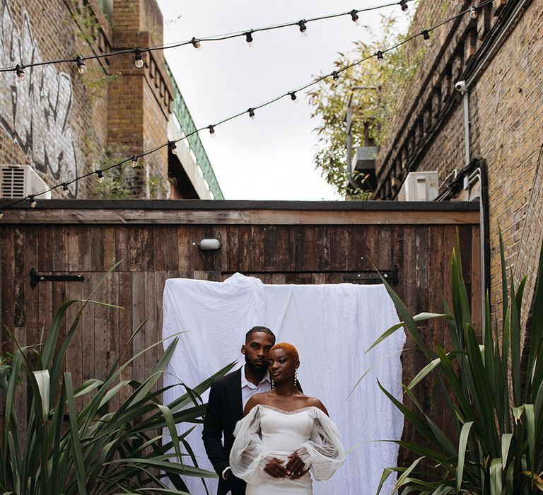 Groom in skinny black suit posing with bride in off shoulder wedding dress with lace puff sleeves standing in front of white drape backdrop at 100 Barrington