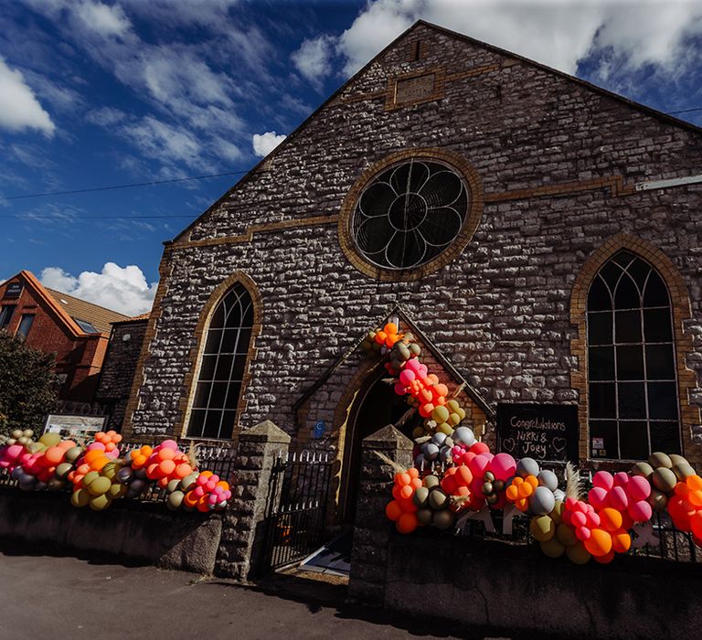 Zion venue in Bristol with pink, orange, olive green, grey and dark grey balloons hung up on the fence with pampas grass floral decorations