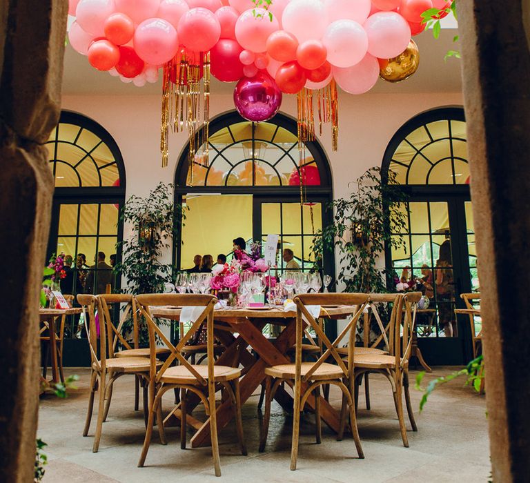 Reception room at Middleton Lodge; wooden tables and chairs, large green foliage decorations and large semicircle of pink hanging balloons as well as large fuchsia disco ball decoration