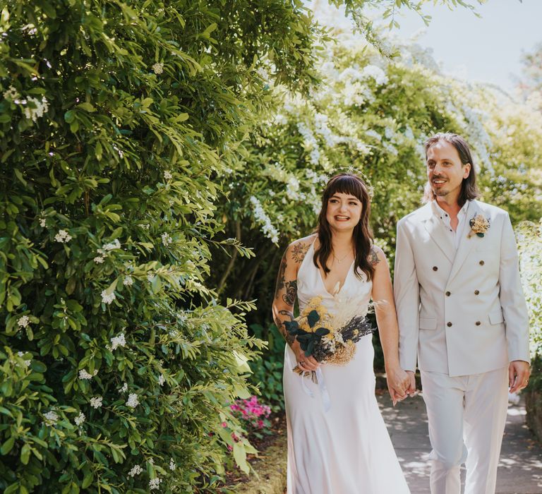 Bride in silk-look v-neck sleeveless ASOS wedding dress and gold halo headband with sparkly stars holding dried flower wedding bouquet with white bunny tails, green foliage, yellow dried gypsophila, lavender and baby’s-breath and groom in neutral coloured double-breasted ASOS suit