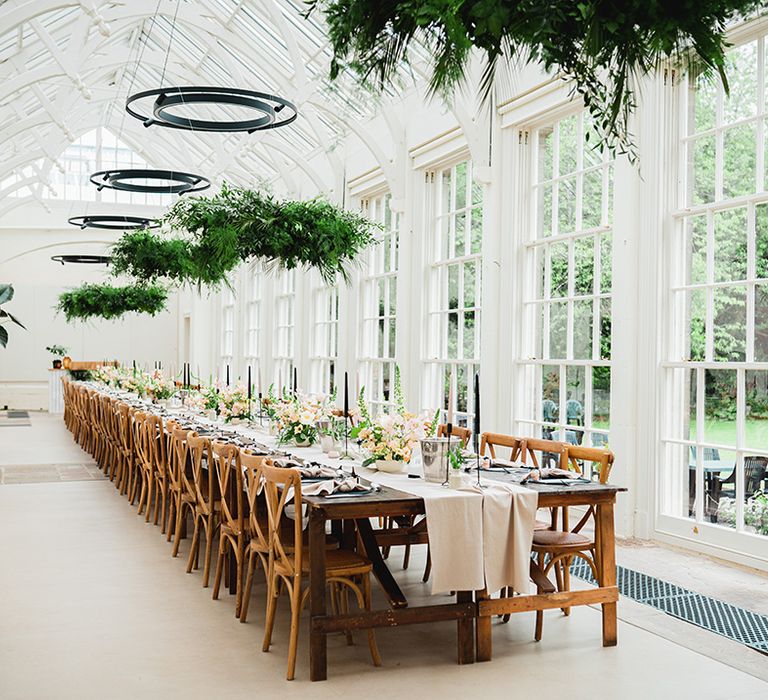 The Orangery Ingestre reception room, with large windows, glasshouse venue roof, festoon chandelier lighting instalments, suspended foliage decorations and neutral, botanical wedding tablescape theme