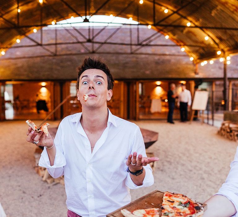 Groom in white shirt and pink corduroy trousers eating pizza at boho wedding