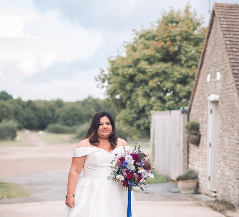 Bride in an off the shoulder wedding dress with blue, purple, and red with foliage tied with blue ribbon 