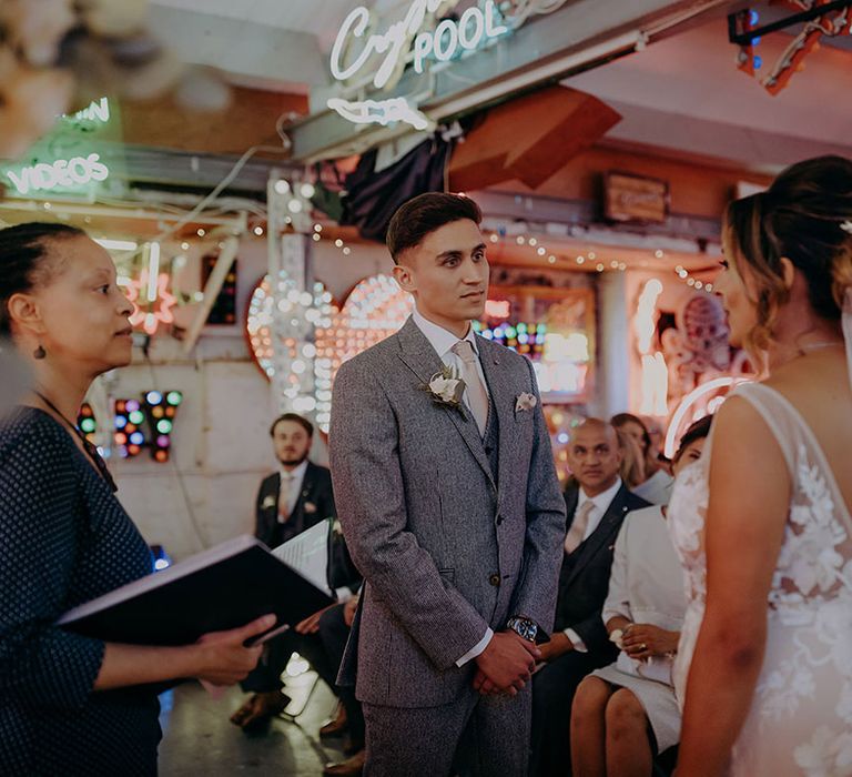 Bride and groom standing at the alter of Gods Own Junkyard with neon signs, LED signs, and fairy lights in the background 