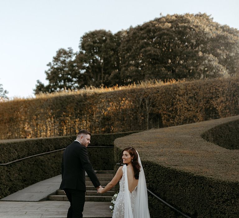 Groom in black suit holding hands with the bride in an open back fitted wedding dress enjoying the gardens at the venue  