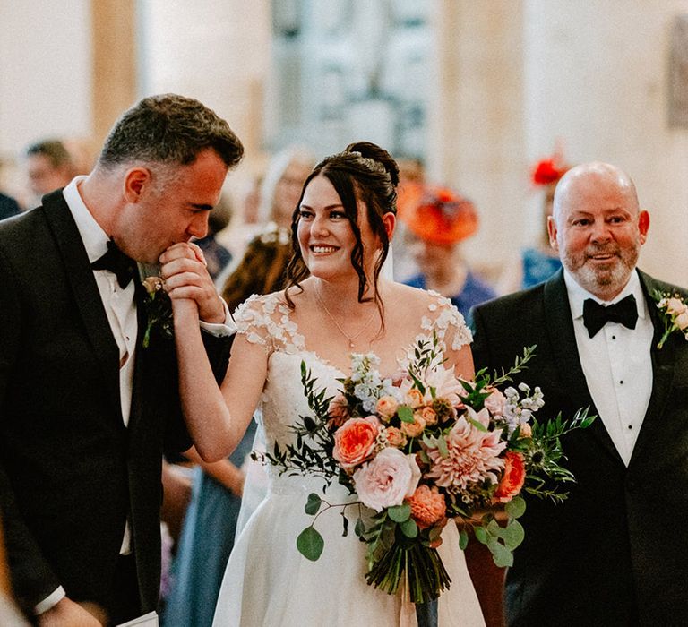 Groom wearing a black tuxedo kisses the bride's hand as she walks down the aisle to meet him for the start of the church wedding ceremony 