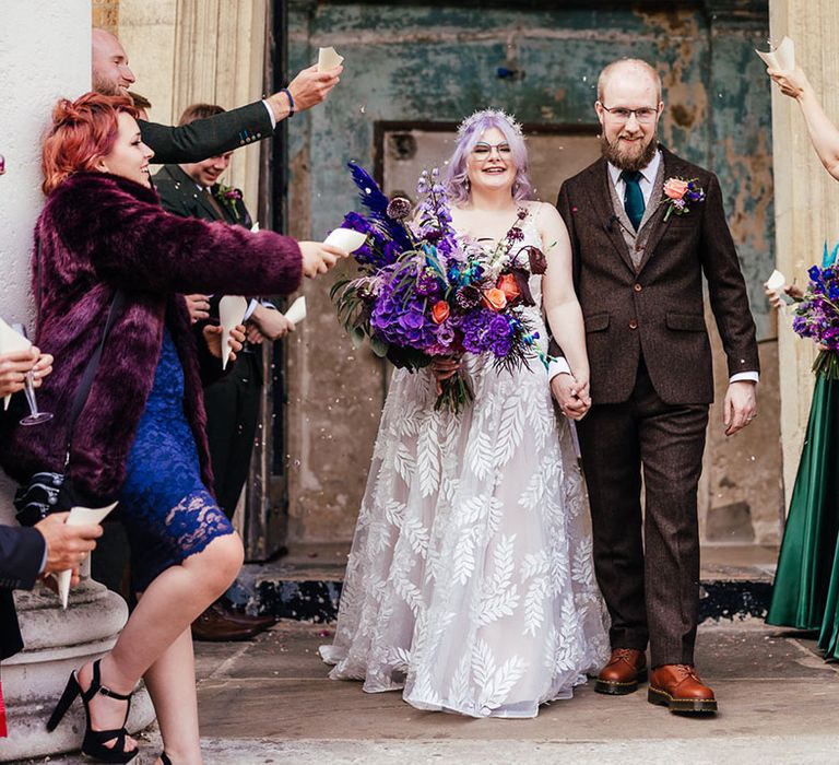 Bride wearing leaf appliqué sheer bridal gown and cathedral length veil holding bright purple bridal bouquet with purple hydrangeas, pink garden roses, purple orchids, lilac carnations, dried flowers and peacock blue feathers and holding hands with groom in mocha brown textured suit with deep green tie and pink and purple boutonniere doing confetti exit at London wedding 