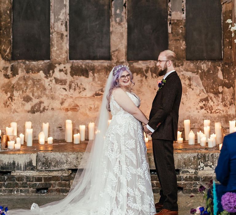 Groom in mocha brown textured suit with pink and purple boutonniere holding hands with bride  in dainty leaf 3d appliqué sheer bridal gown and cathedral length veil at Asylum Chapel London wedding venue with pillar candles and peacock wedding aisle flower arrangements 