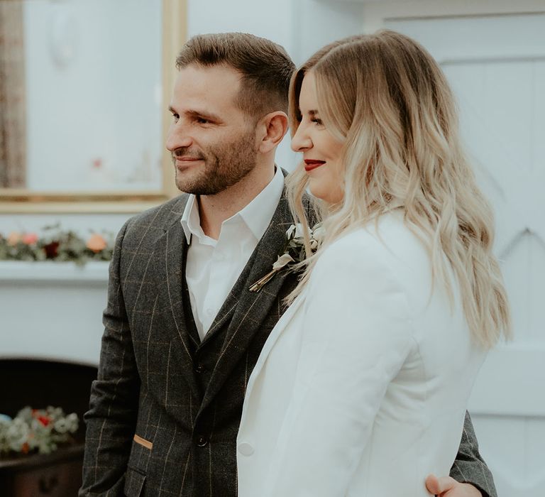 Groom in dark grey checkered suit stands with his arm around the bride wearing a plain silver wedding band 