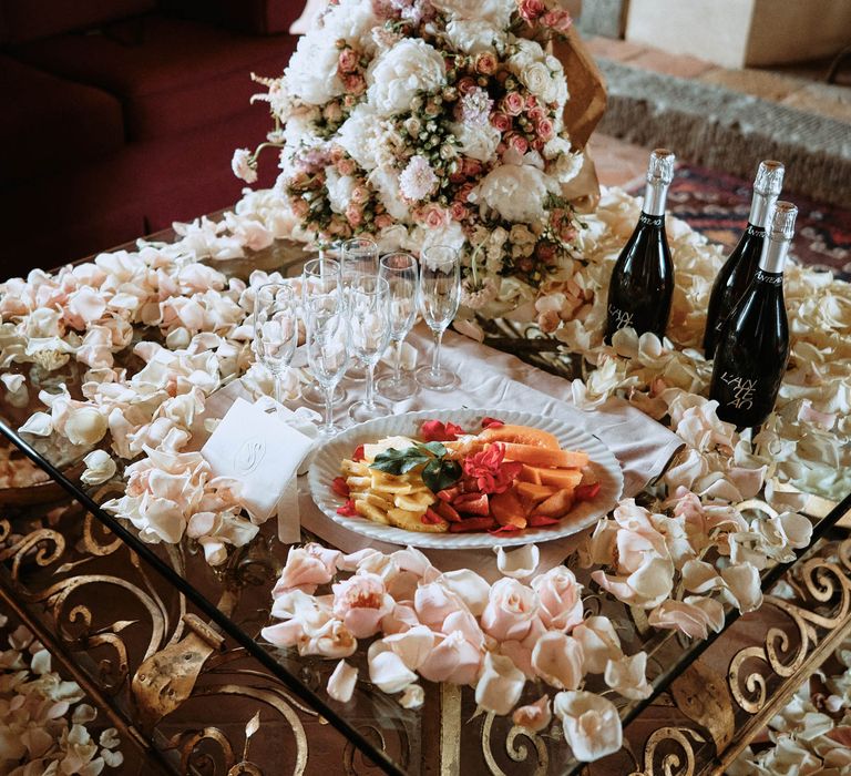 Fruit platter and prosecco surroundeed by pastel pink petals and white roses on the morning off the wedding