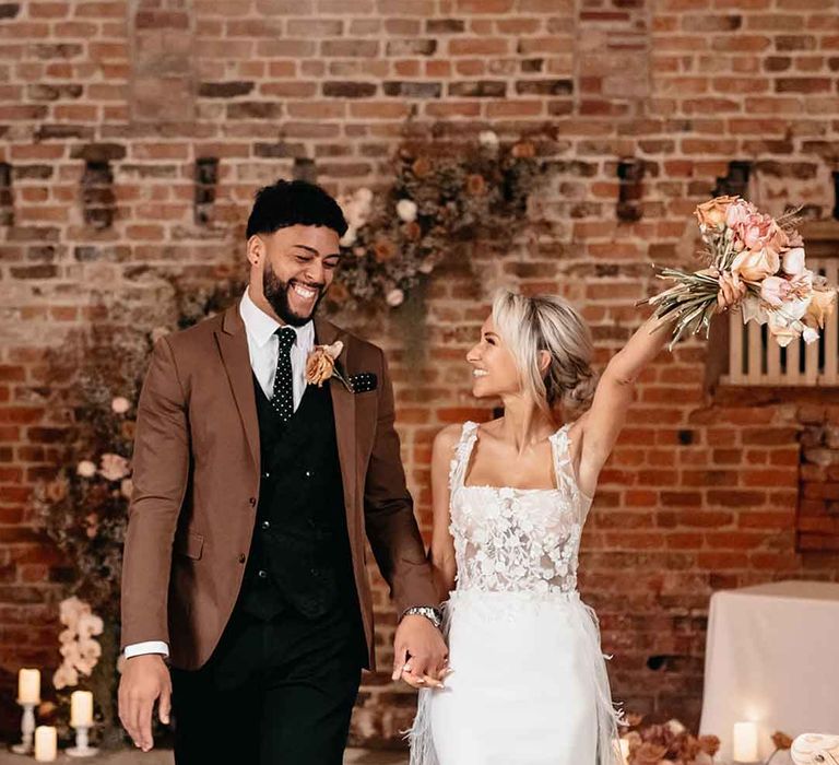 Bride in corset top sheer floral 3d appliqué wedding dress with fringe overlay holding up dried blush garden rose, peony and dried flower bridal bouquet walking with groom in mocha brown blazer, black waistcoat, polka dot tie and pocket square and dried blush boutonniere at Lodge Farm wedding venue 