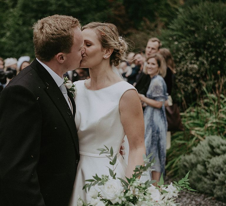 Groom in navy wedding suit with the bride in a high neck wedding dress sharing a kiss after their confetti moment 