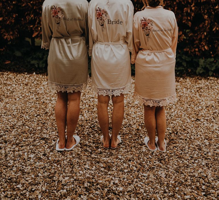 Bride, mother of the bride, and maid of honour in matching satin robes in different colours with their wedding role on the back 