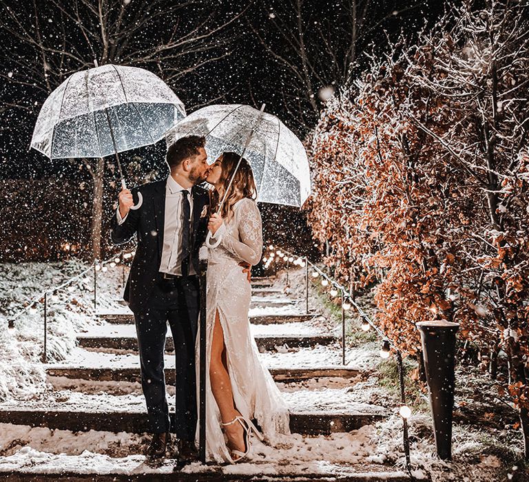 The bride in her sparkly wedding dress kisses the groom in his navy suit under clear umbrellas on their snowy November wedding