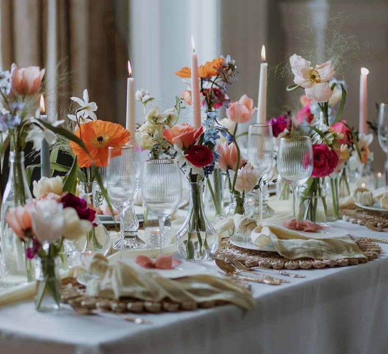 Spring themed wedding tablescape with orange poppies, pink and red roses, jasmine and bluebells on white wedding tablecloth and off-white satin table runner with pastel tapered candles, gold-rimmed wine glasses and classic crockery 