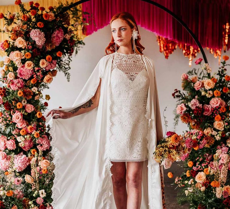 Bride wearing 70s-inspired boatneck short lace wedding dress with detachable tulle puff sleeves and platform gold wedding heels and scallop hem bridal cape standing by circular floral arch with vibrant flowers and foliage attached and pink, red, yellow and metallic wedding streamers behind