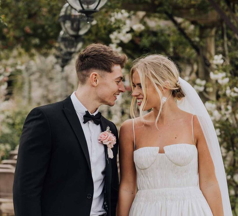 Groom in classic black tuxedo with bowtie and blush rose boutonniere holding hands with bride in Watters strappy sleeveless princess wedding dress and chapel length wedding veil 