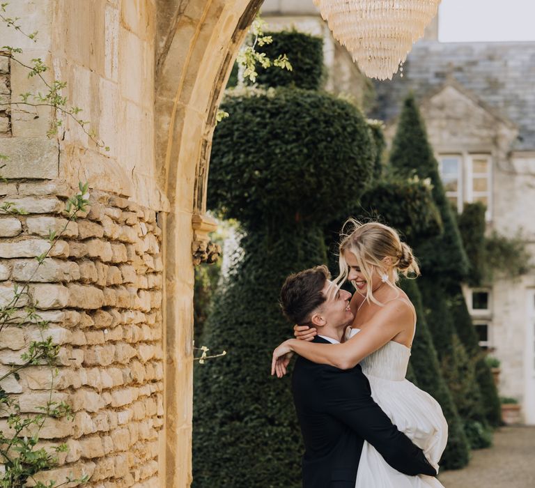 Groom in classic black tuxedo picking up bride in Watters strappy sleeveless princess wedding dress under crystal chandelier at Euridge Manor wedding