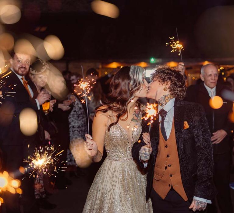 Bride in dark velvet bridal jacket with rust waistcoat and pocket square, black suit trousers and gold pocket watch kissing bride in sleeveless deep v-neck sparkly gold wedding dress doing wedding sparkler send-off at Kingscote Barn Tetbury LGBTQIA+ wedding