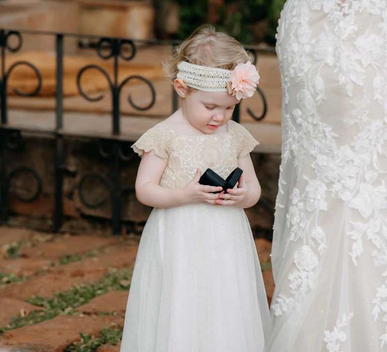 Child at intimate destination elopement wedding at Giraffe Manor wedding venue in Kenya wearing off-white dress and sequin headband with pink carnation holding wedding ring box