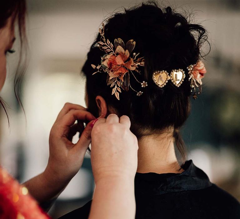 Bride with dark brown wedding hair being secured into an updo with flower hair accessories and personalised gold and pearl heart hair pins 