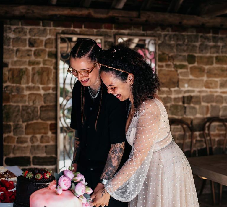 Bride in silver chains, glasses, black shirt and black trousers cutting wedding cake with bride in long sleeve v-neck sheer polka dot overlay sparkly wedding dress and gold celestial bridal headband 