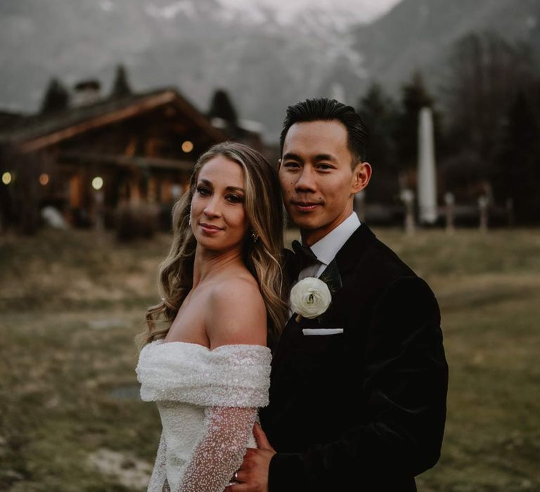 Bride in off shoulder sparkly mermaid wedding dress standing with groom in classic black tuxedo, black bowtie and white garden rose boutonniere at French destination ski wedding