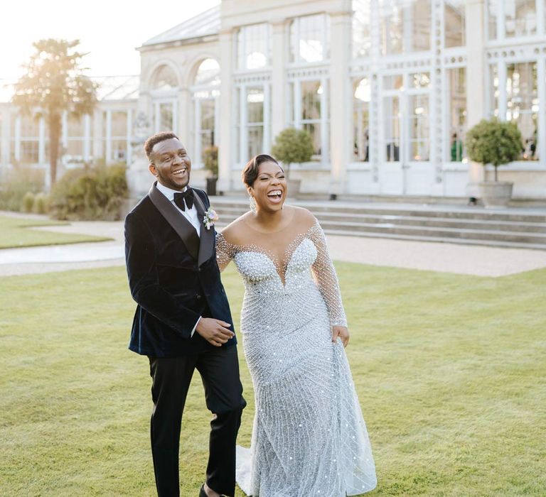 Bride in illusion v-neck sheer off shoulder long sleeve pearl embellished wedding gown laughing with groom in velvet midnight blue double breasted groom’s blazer, velvet bowtie and peach garden rose boutonniere during vow renewal at Syon Park wedding venue