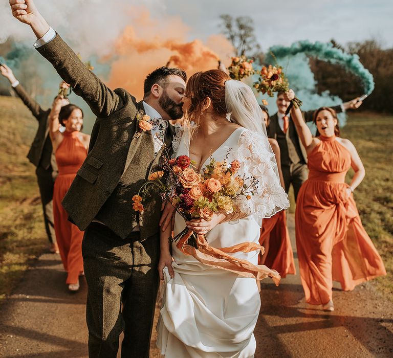 Groom in tweed three piece wedding suit kissing bride in Suzanne Neville wedding dress with bridesmaids in orange bridesmaid dresses 