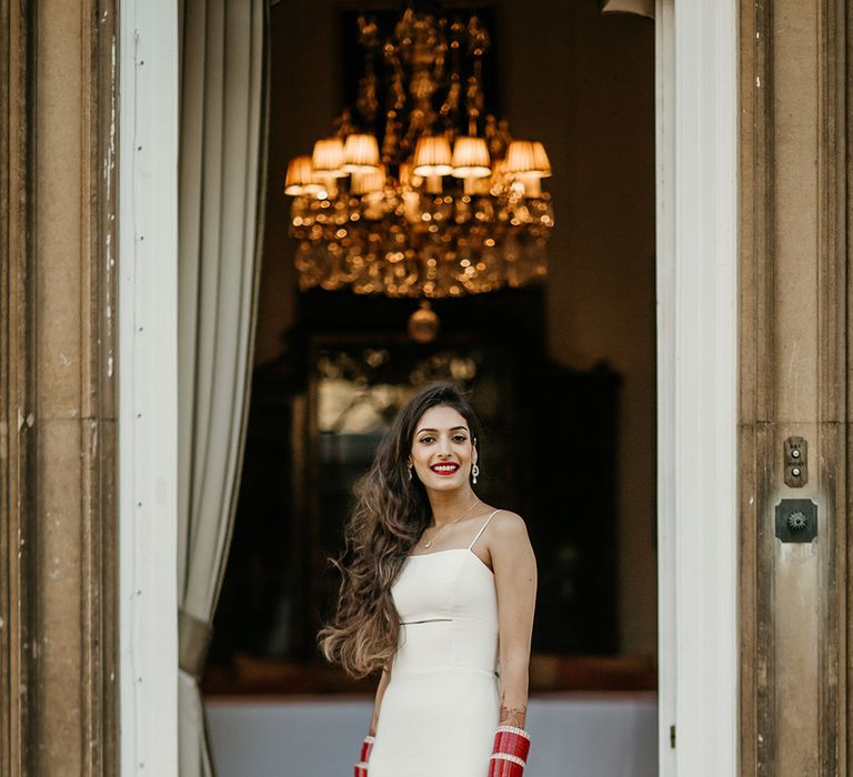 Bride with brown tousled wedding hair in fitted slip wedding dress with midriff cutout wearing red bangles 