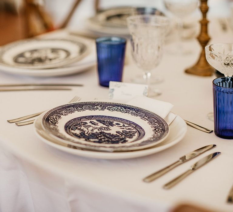 Patterned china plates with blue coloured glassware and white table cloth 