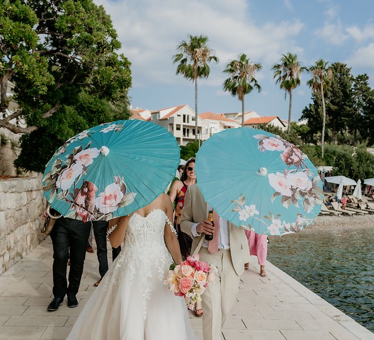 Bride in 3D lace wedding dress and groom in linen suit walk along the side of the ocean with wedding guests following while holding turqouise South Asian umbrellas