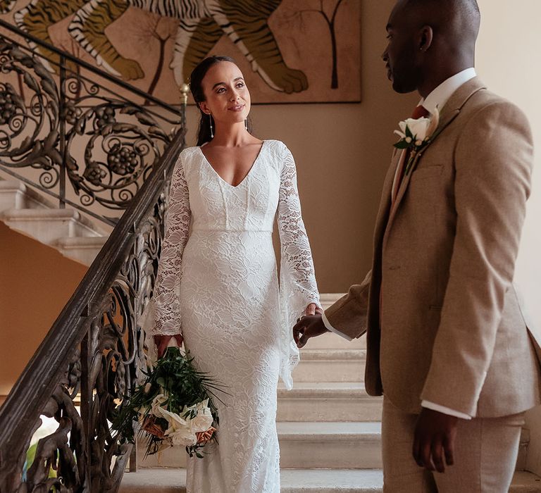 Bride walking down stairs wearing a long sleeve boho lace wedding dress with the groom in a beige suit