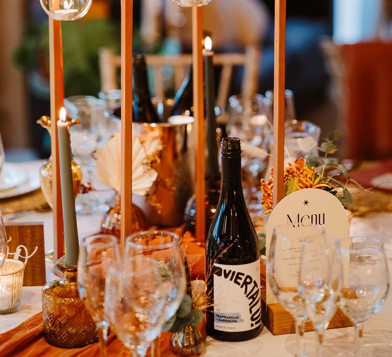 Orange autumnal wedding table centrepiece with orange and green candles, wedding menu sign and bottles of alcohol 