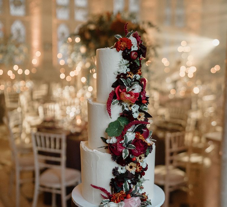 Three tier white iced wedding cake decorated with flowers lining one side of the cake with deep red, orange and light pink tones 