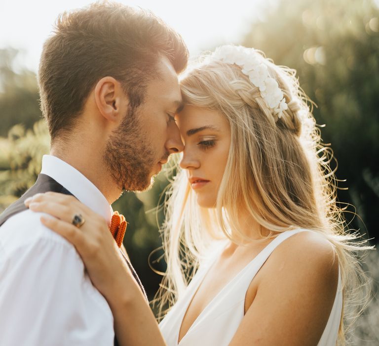 Bride and groom embrace as the sunlight illuminates the bride's golden hair and stunning white flower headband