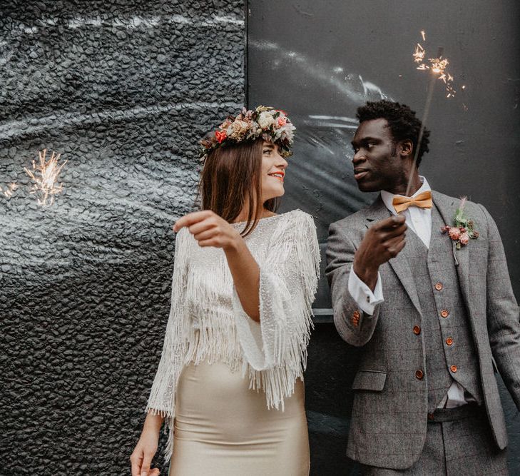 White bride in tassel dress and Black groom in tweed suit both hold sparklers and look at eachother