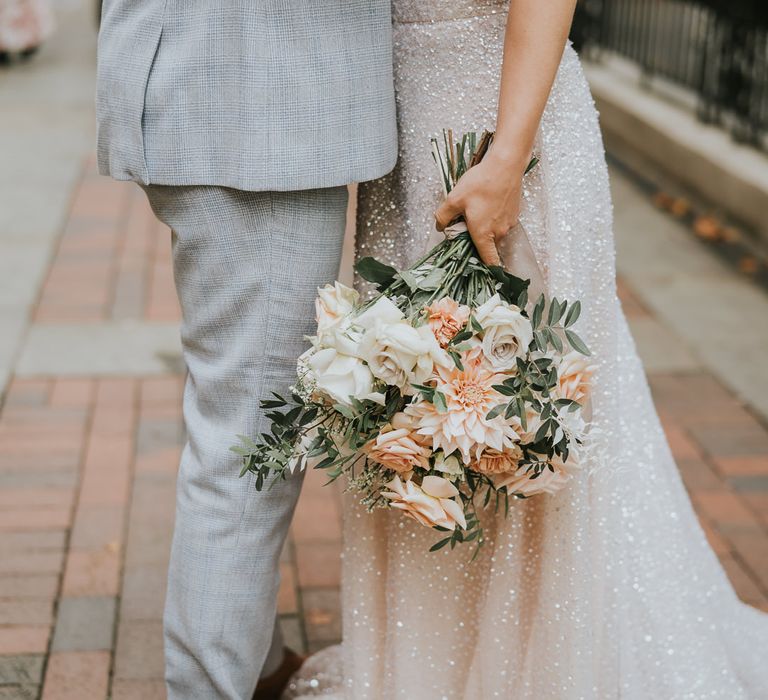 Groom in a grey wedding suit with blue check detail standing neck to his bride in sparkly separates holding a blush pink, peach and white bouquet with roses and dahlias 