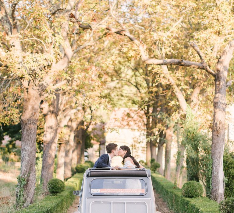 A vintage wedding car drives off with tin cans attached to the back. A couple have their heads out of the sun roof and kiss. Image by Hannah Duffy Photography./