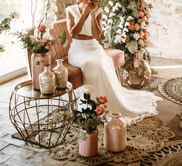 Bride in separates sitting on a velvet sofa surrounded by boho wedding flowers and decor 