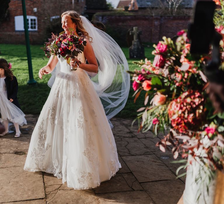 Bride laughs whilst holding bouquet and veil blows in the wind