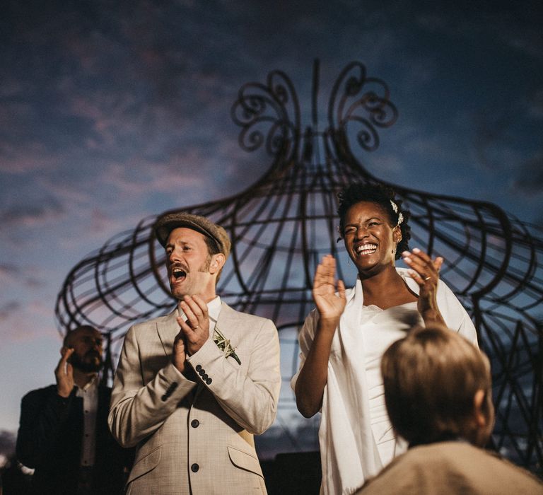 Groom in a beige Peaky Blinder wedding suit and flat cap clapping at the outdoor wedding ceremony with his bride 