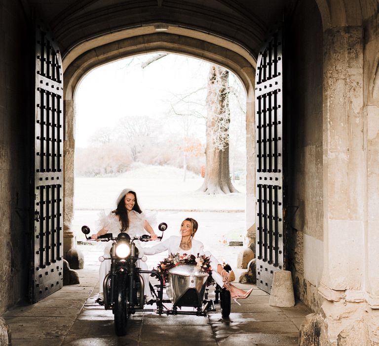 Two brides sitting on a motorcycle and wedding sidecar at the grand entrance to Ashridge House in Herfordshire 