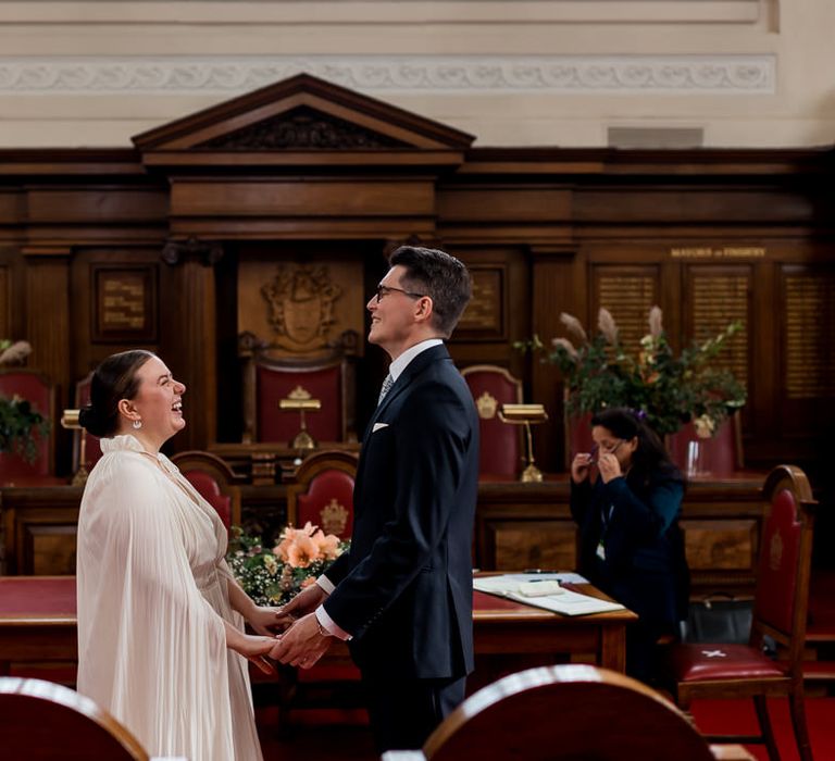 Groom in a navy suit and bride in a bridal cape holding hands at the altar in Islington Town Hall