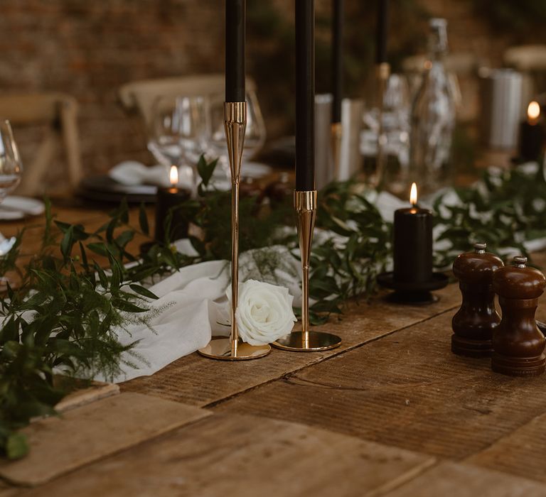 Tall black candles in golden candlesticks for rustic wedding table decor with greenery and white rose details