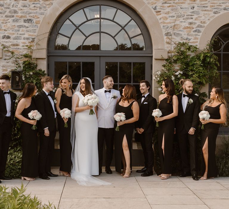 Bride in white Made With Love wedding dress poses outside with groom in white tuxedo jacket, bridesmaids in long black bridesmaid dresses and groomsmen in black tuxedos