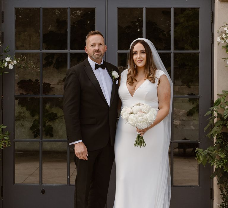 Bride in white Made With Love wedding dress holding white and blush pink rose bouquet poses outside with father in black tuxedo