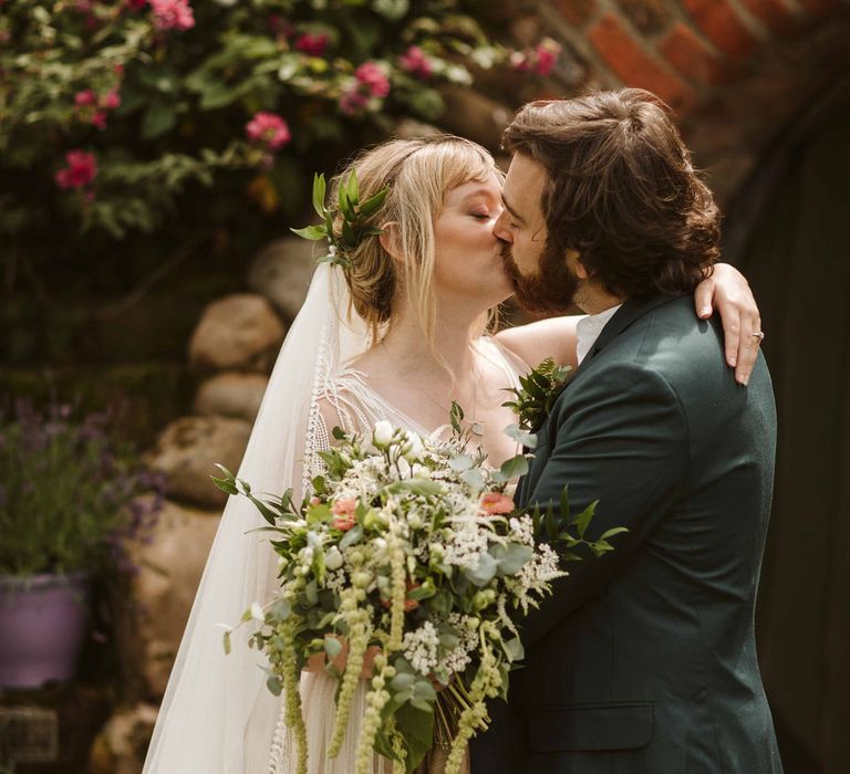 Bride & groom kiss outdoors whilst bride holds white floral bouquet