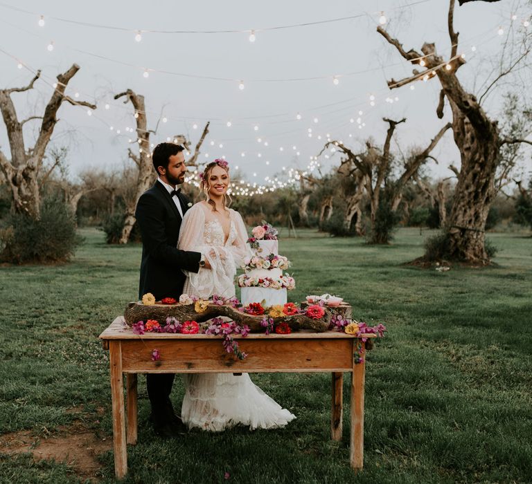 Bride in long sleeved boho wedding dress and groom in navy suit stand hugging at wooden table situated in olive grove, with three tier wedding cake and floral decorations at enchanted forest wedding in Italy